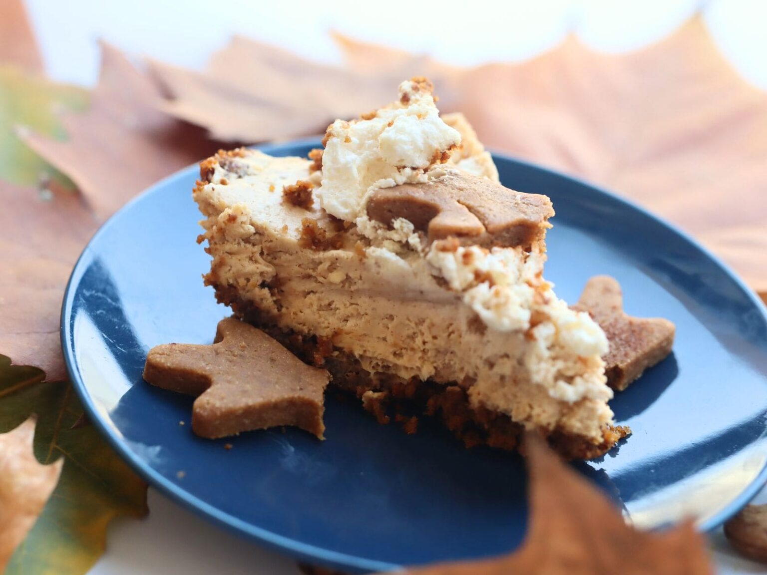 A slice of cheesecake with a crumbly topping and shaped cookie decorations sits on a dark blue plate, surrounded by autumn leaves. The dessert has a creamy texture and hints of spice, creating a cozy autumnal atmosphere.